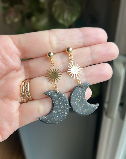 Hand holding two pairs of earrings with moon and sun designs.
