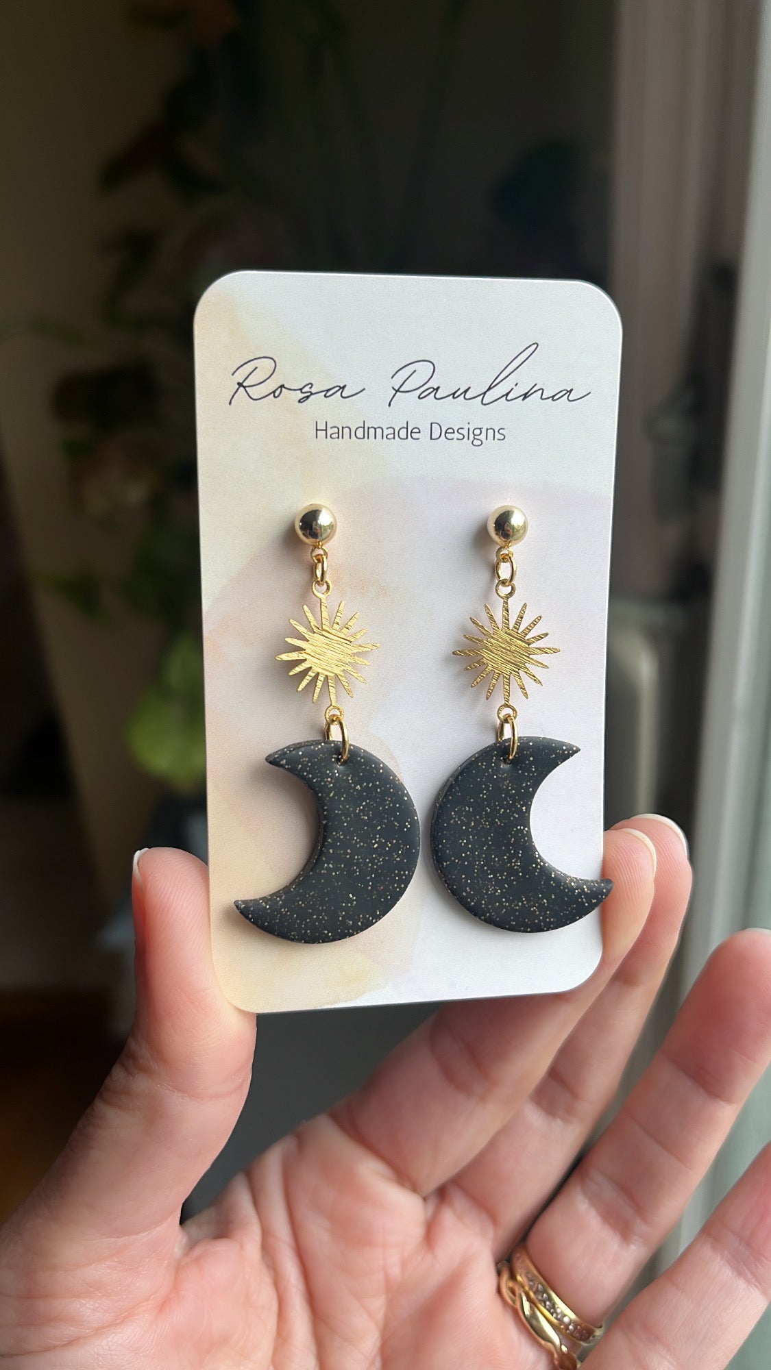 Black and gold earrings on a 'Rosa Paulina' branded card held in a hand.